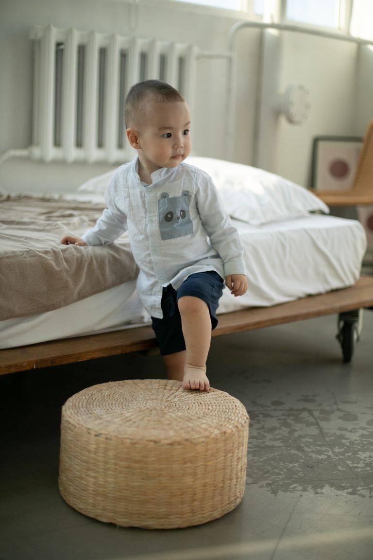 A small child exploring a modern bedroom, stepping on a woven ottoman.