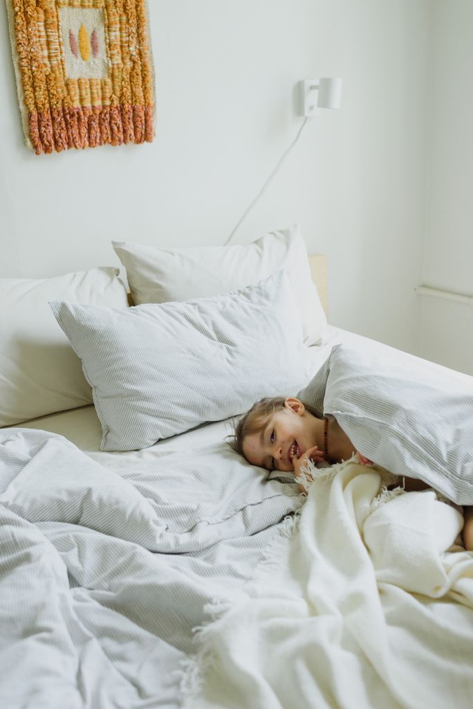 From above of happy curious little kid lying under blanket on bed and smiling in sunny morning